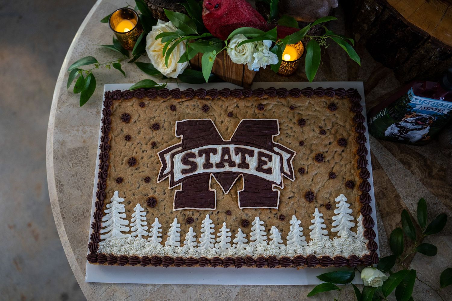 Mississippi State University themed cookie cake with maroon M State logo, chocolate chips, and white frosting trees.