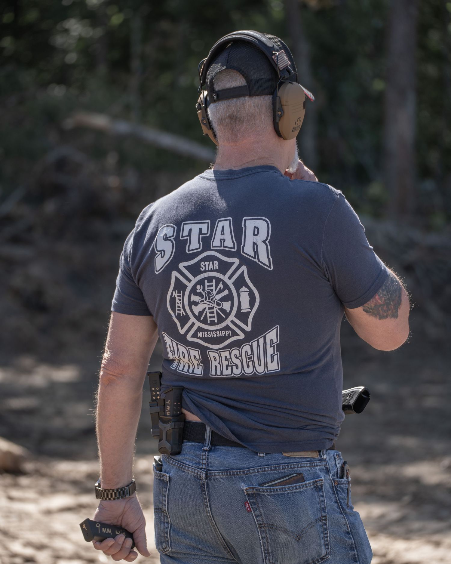 Man wearing Star Fire Rescue t-shirt and ear protection holds firearm at an outdoor shooting range.