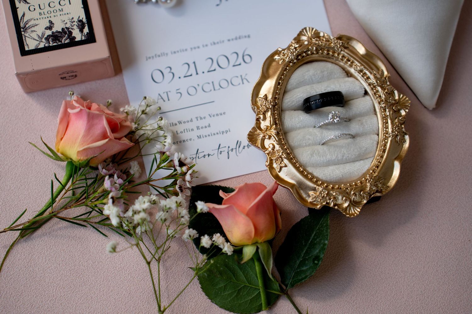 Wedding flat lay with gold ring frame, pink roses, invitation dated 03.21.2026, engagement ring, and Gucci box on blush background.