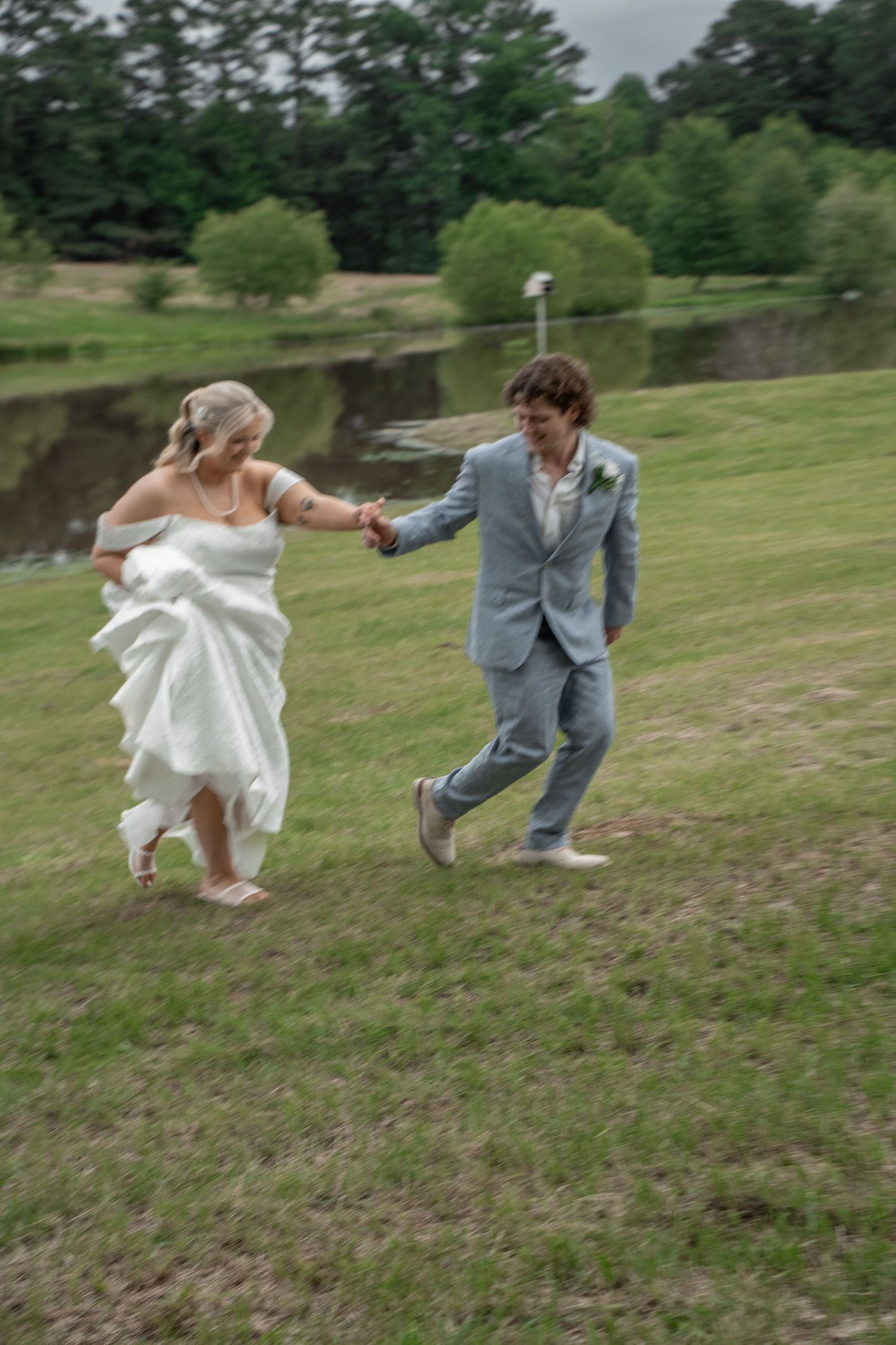A bride in white and groom in gray suit run hand-in-hand across green grass near a pond.