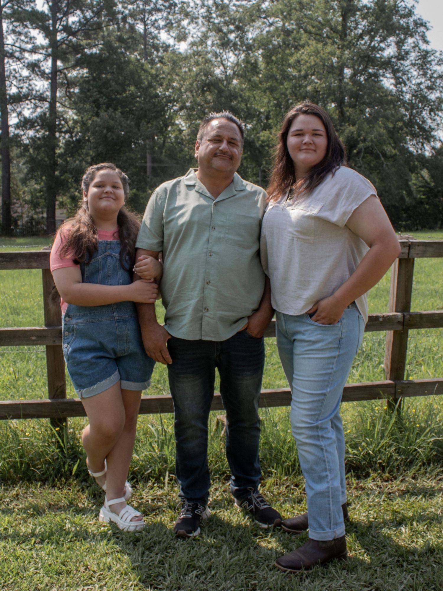 A man and two girls pose smiling by a wooden fence on a green lawn with trees in the background.