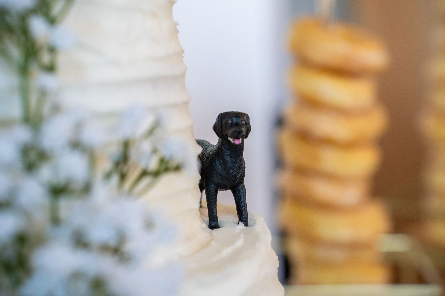 Black Labrador dog figurine topper on white wedding cake with donuts in background.