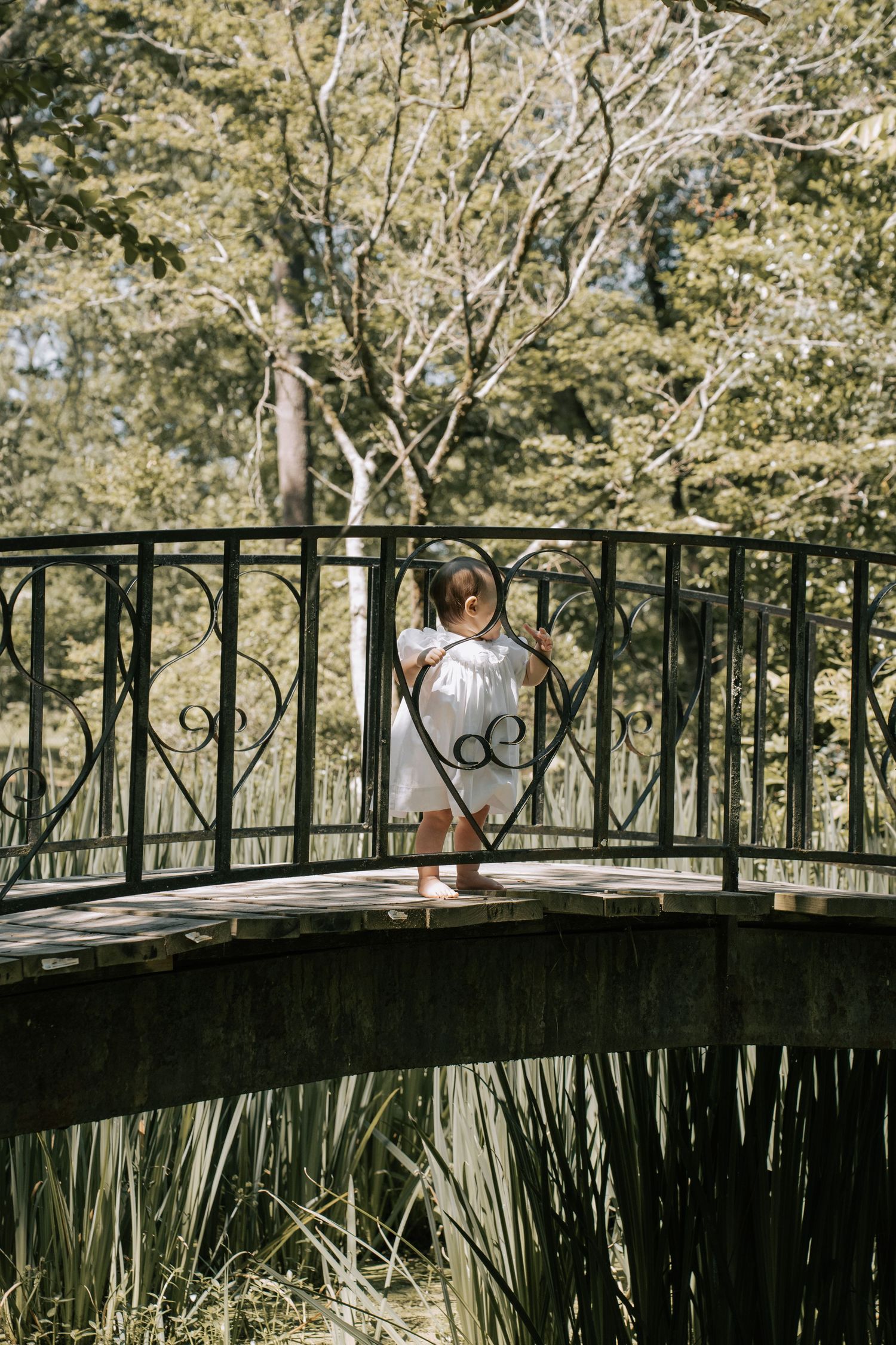 Young girl in white dress standing on ornate iron bridge surrounded by lush green trees in sunlit park.