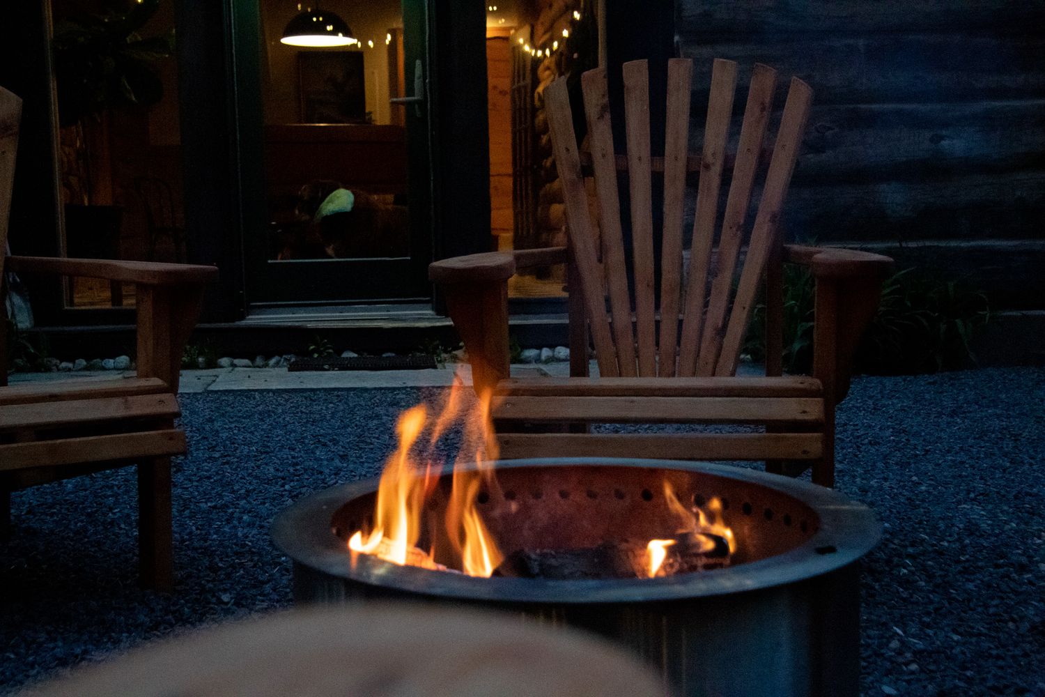 Wooden Adirondack chair beside a glowing fire pit on gravel patio at dusk near a rustic cabin.