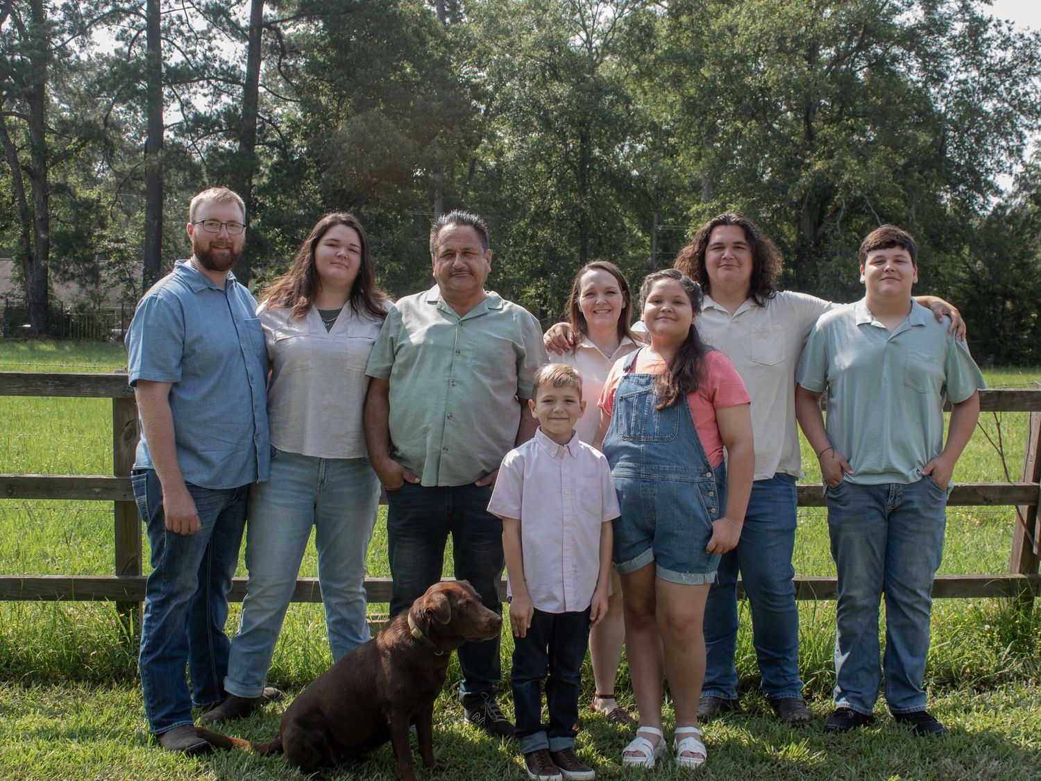 Family group photo outdoors near a wooden fence with green trees, posing with a brown dog on a sunny day.