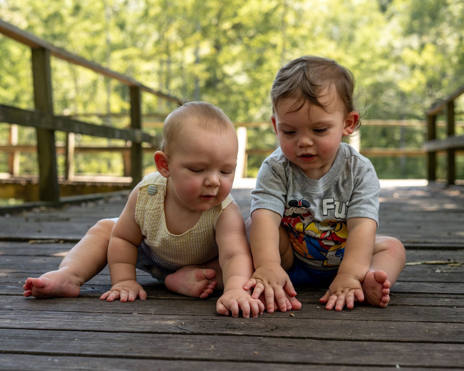 Two babies sitting together on a wooden deck outdoors, exploring with their hands on a sunny day.