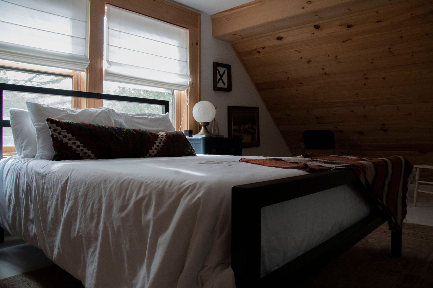 Cozy attic bedroom with white bedding, dark metal bed frame, wood-paneled walls, and natural light from windows.