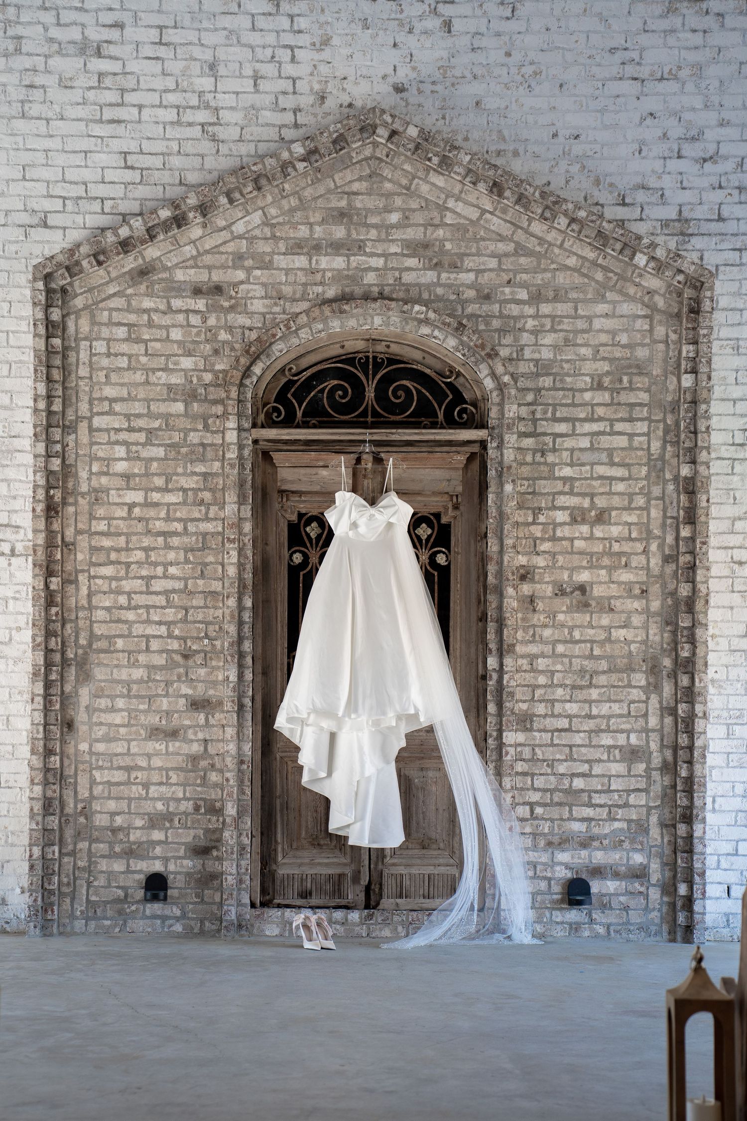 White wedding dress with veil hanging on rustic wooden arched door against whitewashed brick wall.