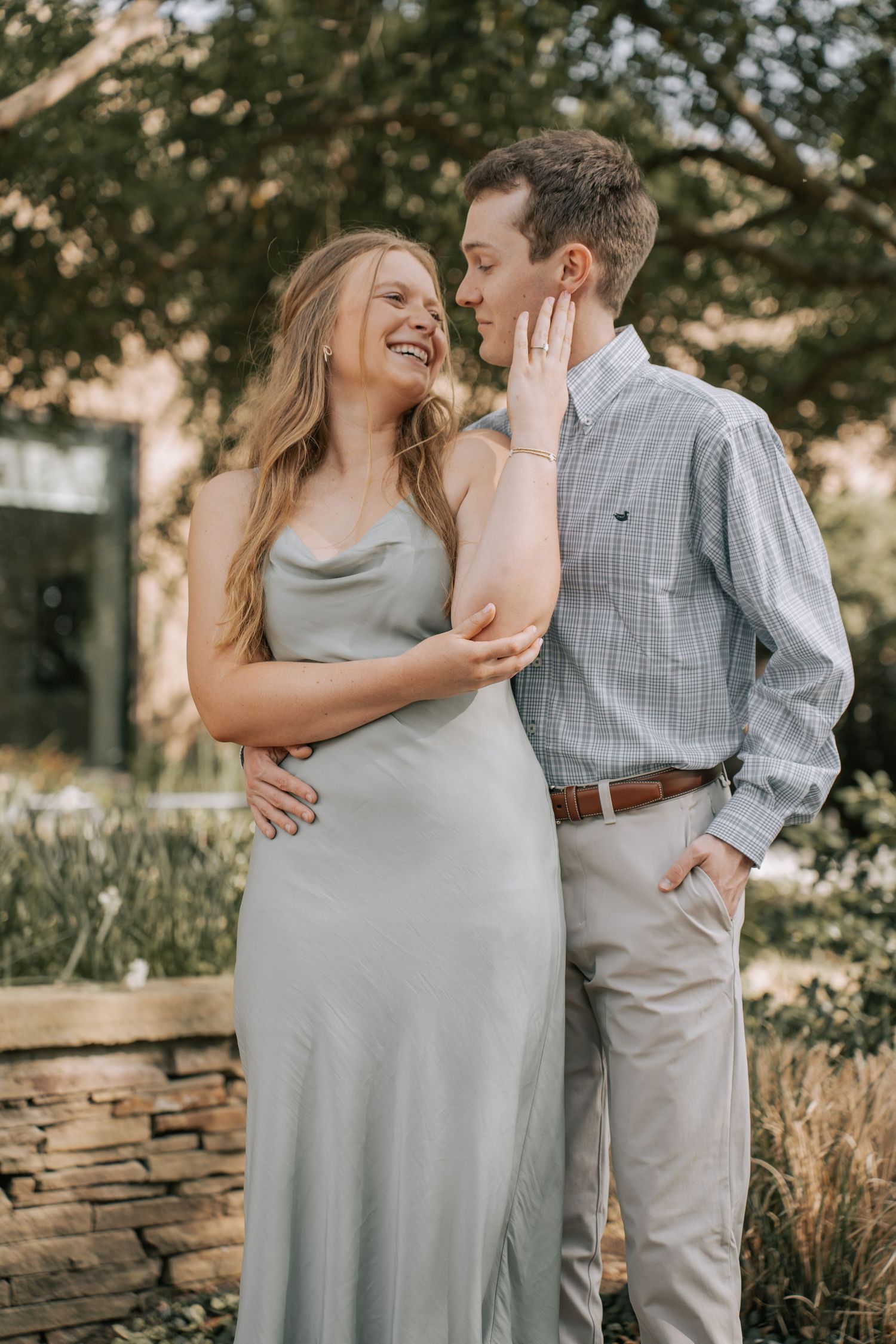 A smiling couple poses outdoors, woman in sage slip dress touches man's face, wearing plaid shirt and khakis.