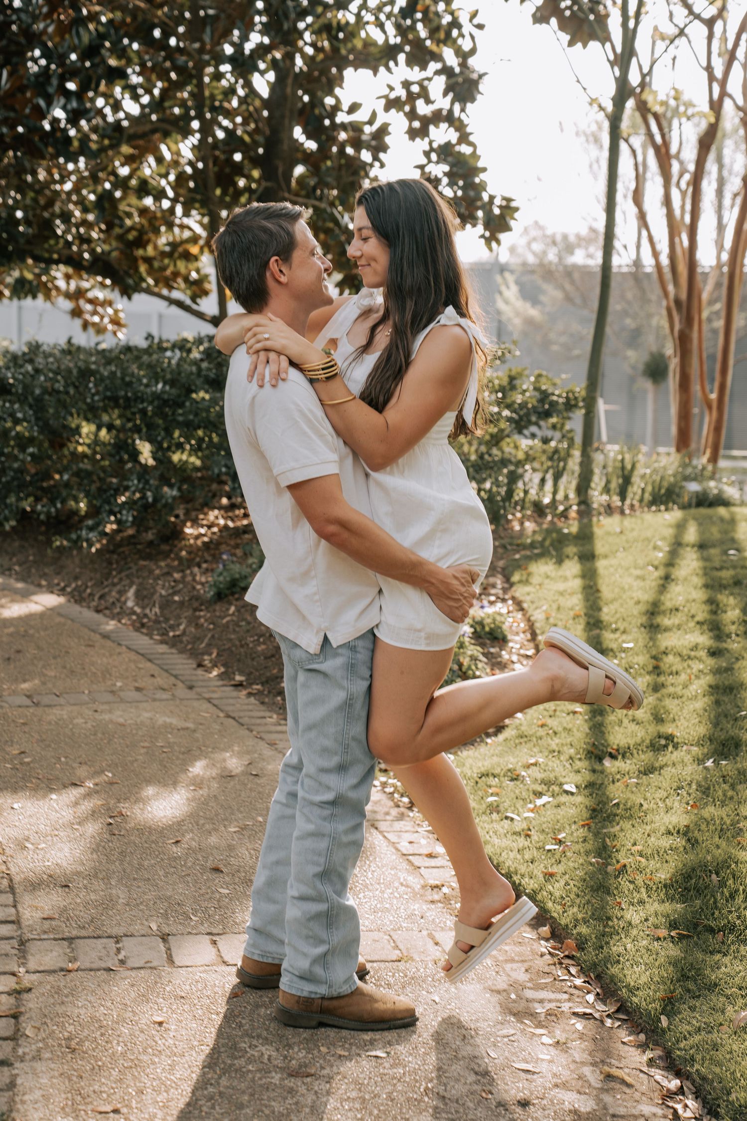 Couple in white outfits sharing a romantic moment outdoors, woman lifting her leg as they embrace on a garden path.