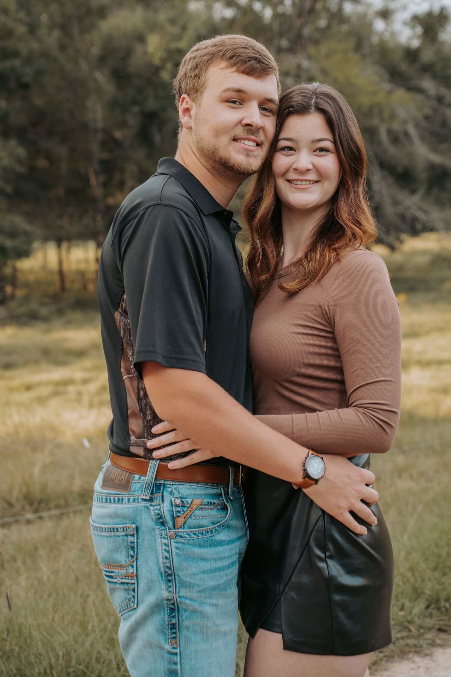 A smiling young couple embraces outdoors in a field, the man in a black polo and jeans, the woman in a brown top and skirt.