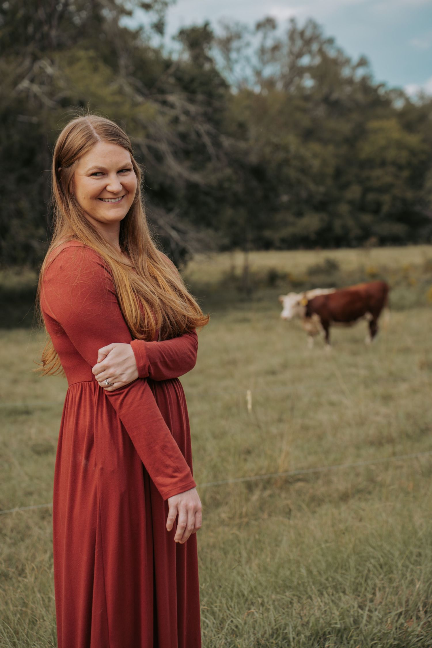 Smiling woman in red maxi dress stands in green pasture with Hereford cow behind her.