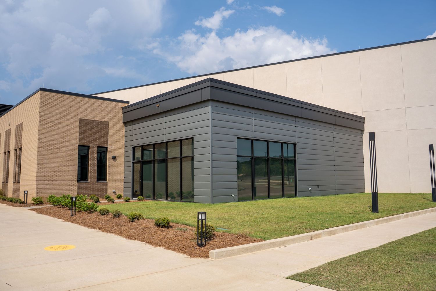 Modern commercial building exterior with gray metal siding, large windows, brick accents, and manicured lawn on a sunny day.