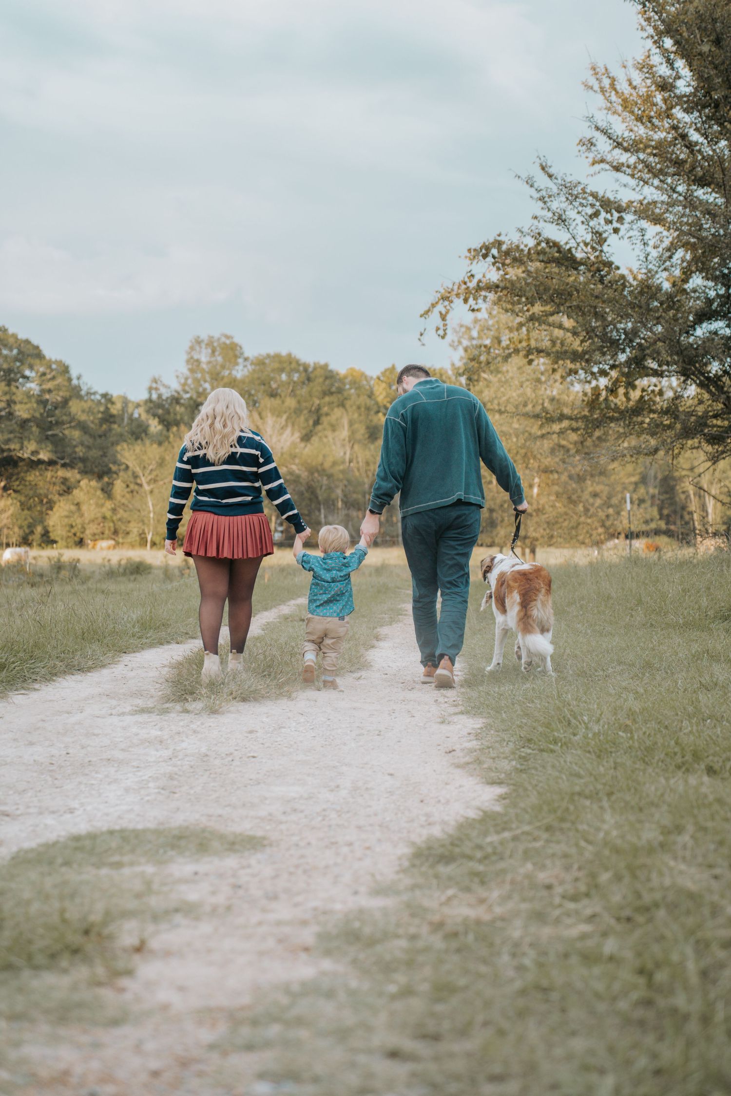 Family with toddler and dog walking on rural path surrounded by autumn trees and green fields.