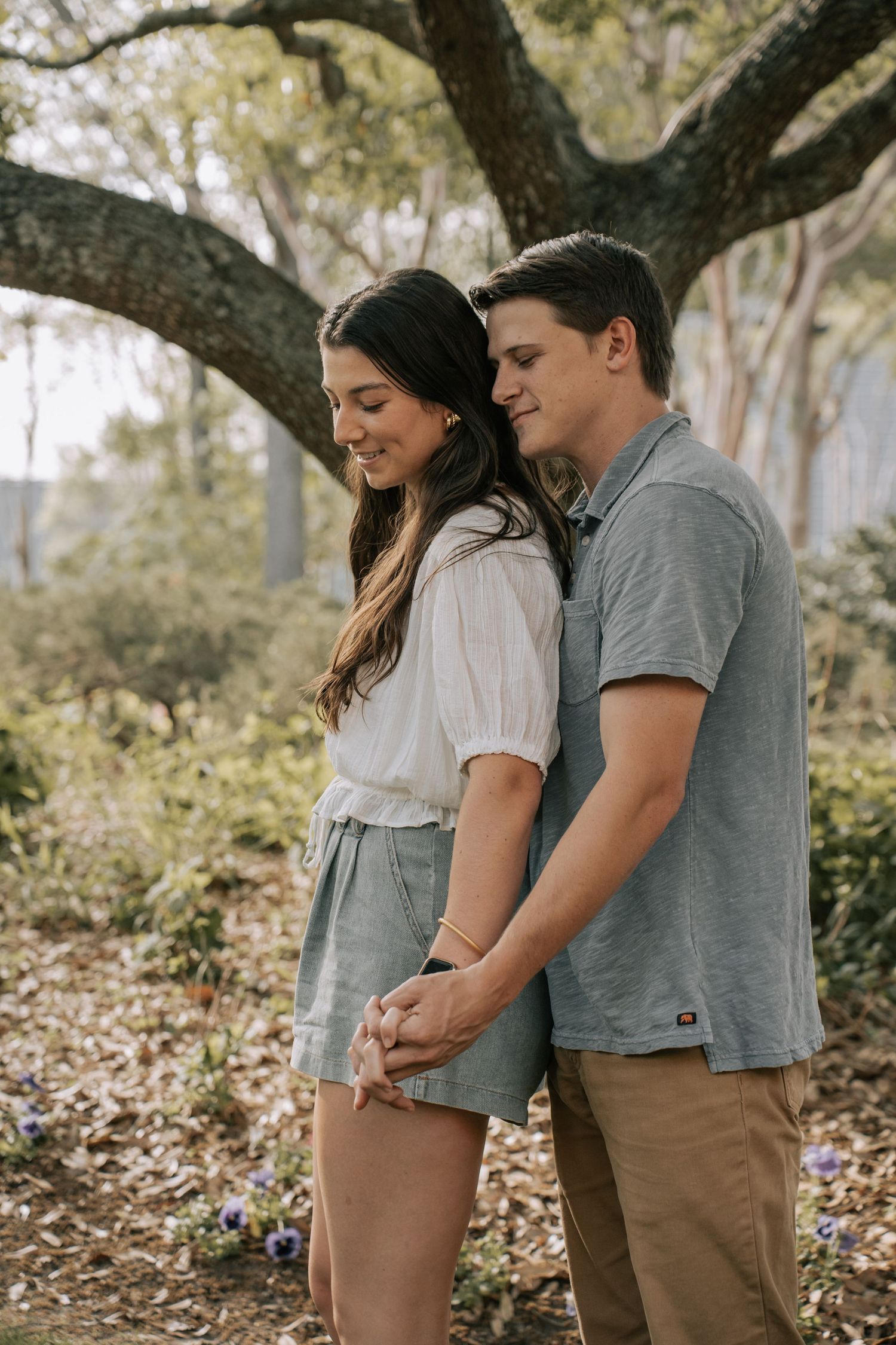 A couple holds hands and smiles while standing together outdoors beneath a large tree surrounded by greenery.