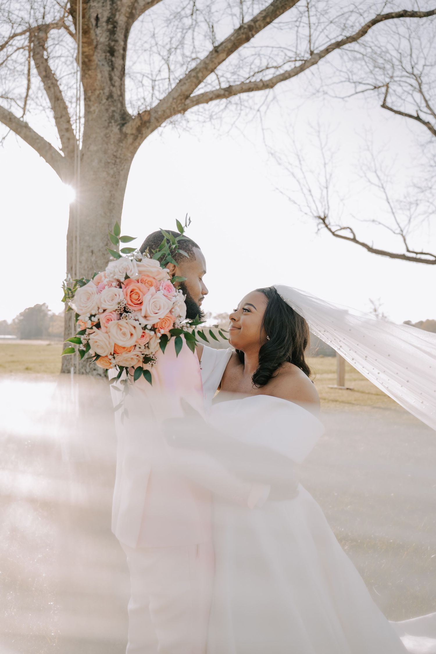 Bride and groom share a romantic moment under a bare tree, with a flowing veil and pink bouquet in soft sunlight.