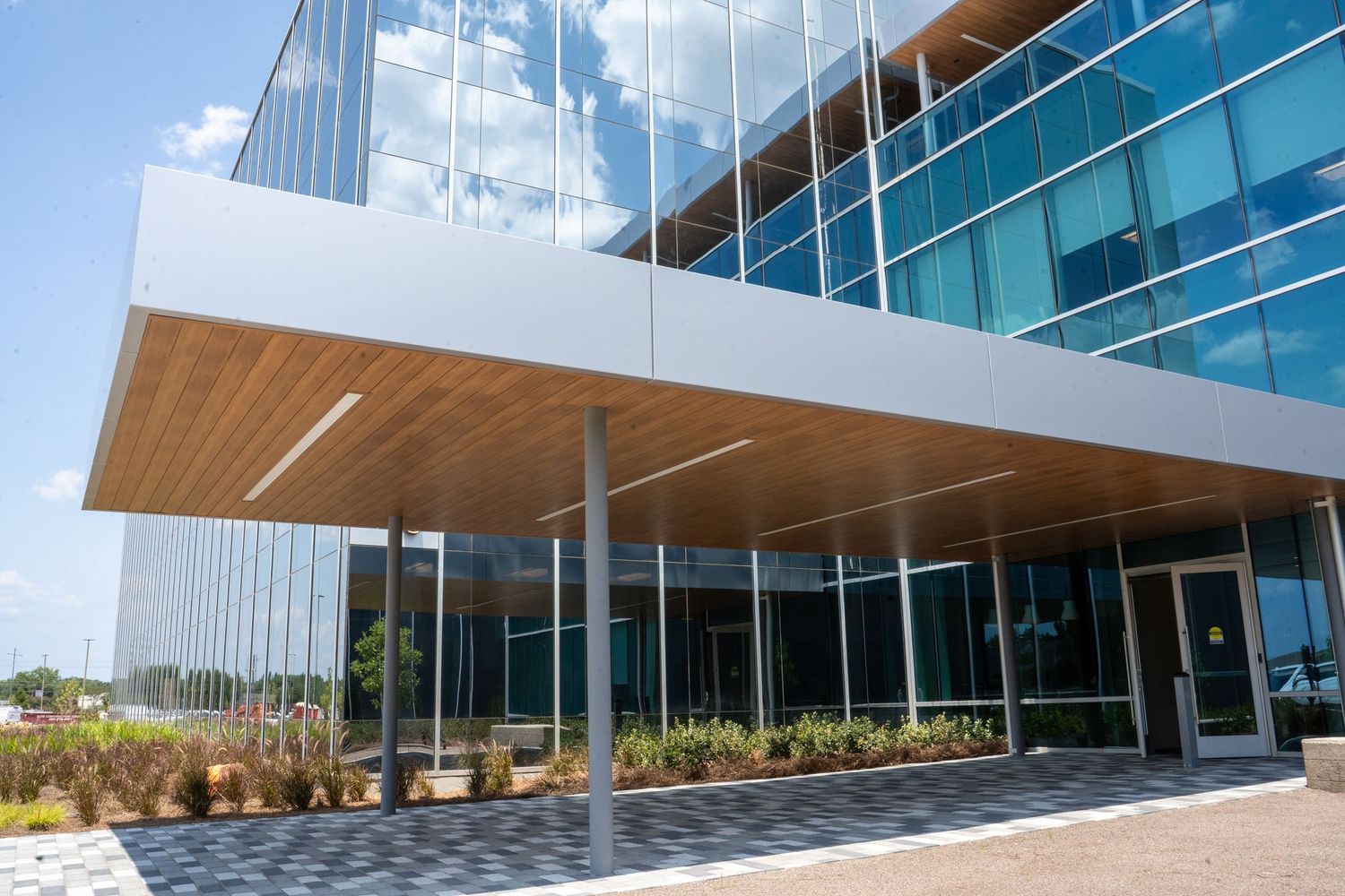 Modern corporate office building with glass facade, wooden canopy entrance, and landscaped walkway under a clear blue sky.