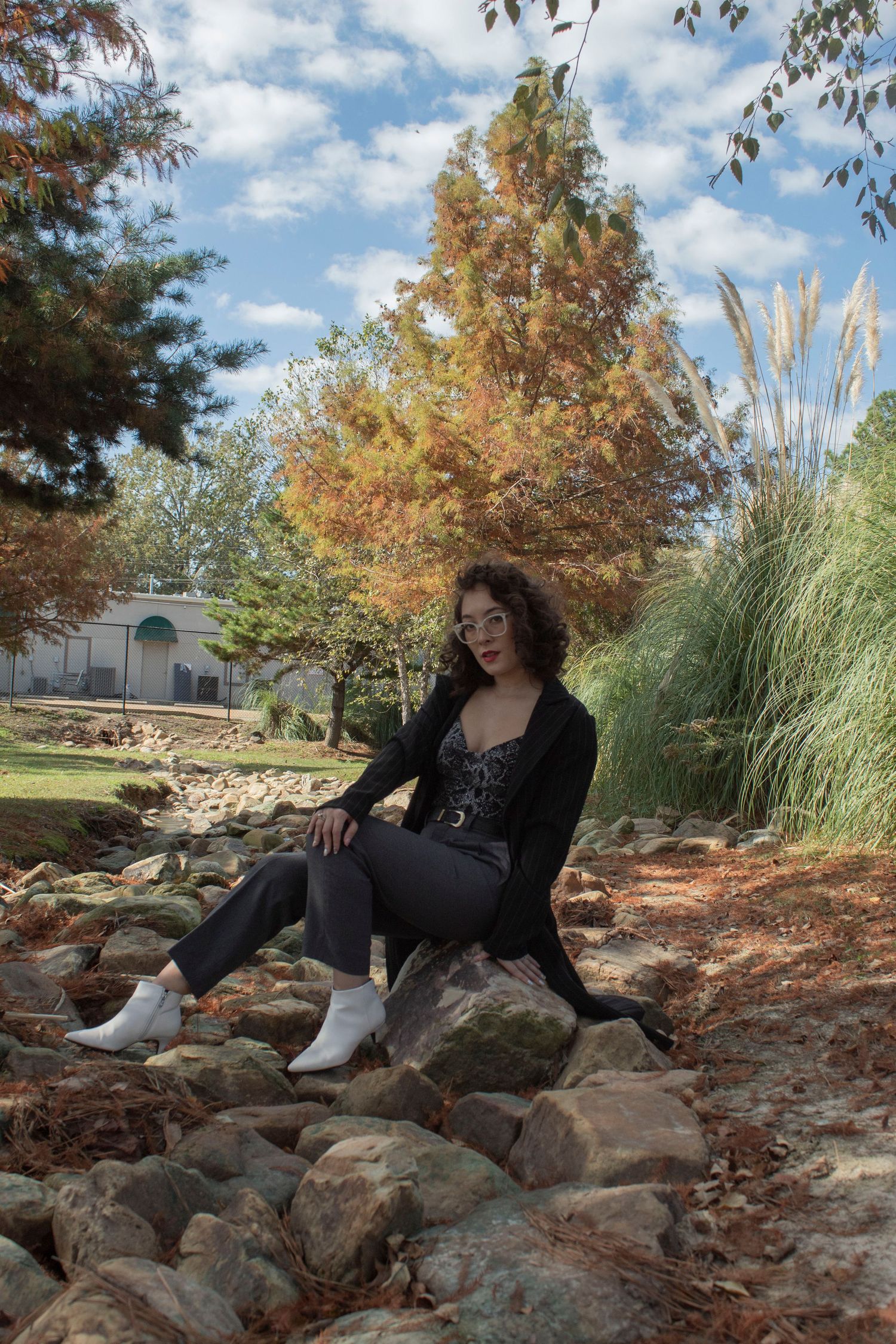 Young woman in black outfit and white boots sitting on rocks in a park with autumn foliage trees.