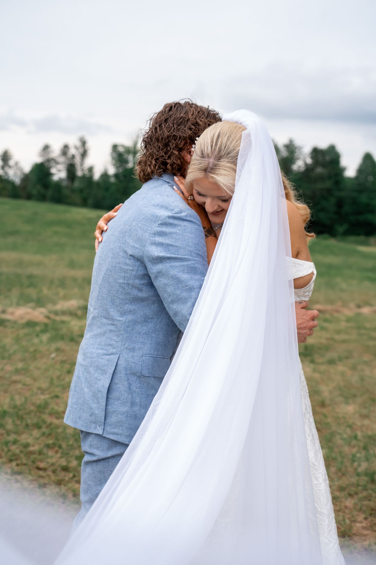 Bride and groom share a tender embrace in a green field, her white veil flowing gracefully as he wears a light blue suit.