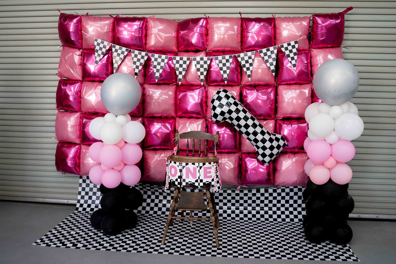 Pink and black checkered party backdrop with hot pink foil balloons, balloon columns, and a small chair for a race car theme.