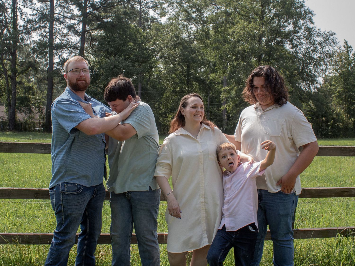 Family of five posing outdoors near a wooden fence with lush green grass and trees in the background.