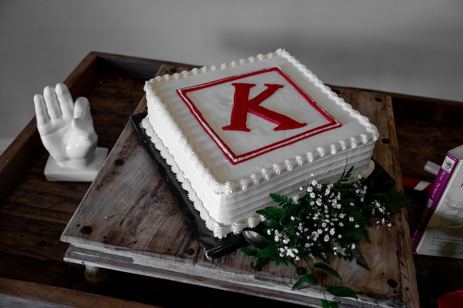 White square celebration cake with red letter K monogram, decorated with white frosting borders and baby's breath flowers on wooden tray.