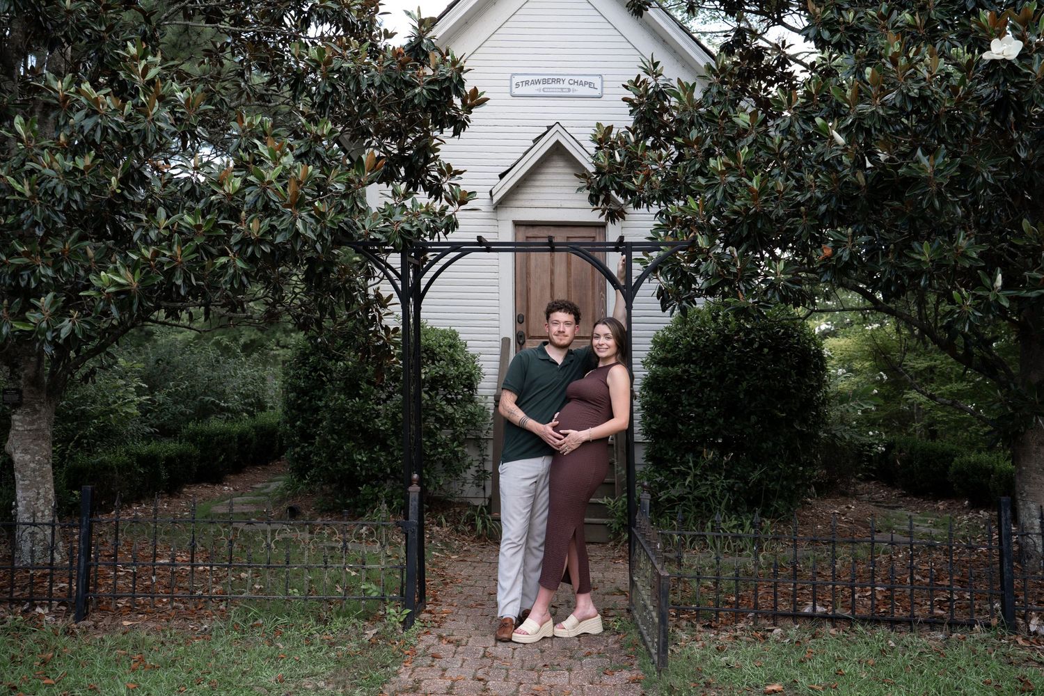 Couple embracing under iron garden gate arch before white chapel surrounded by lush green trees and shrubs.