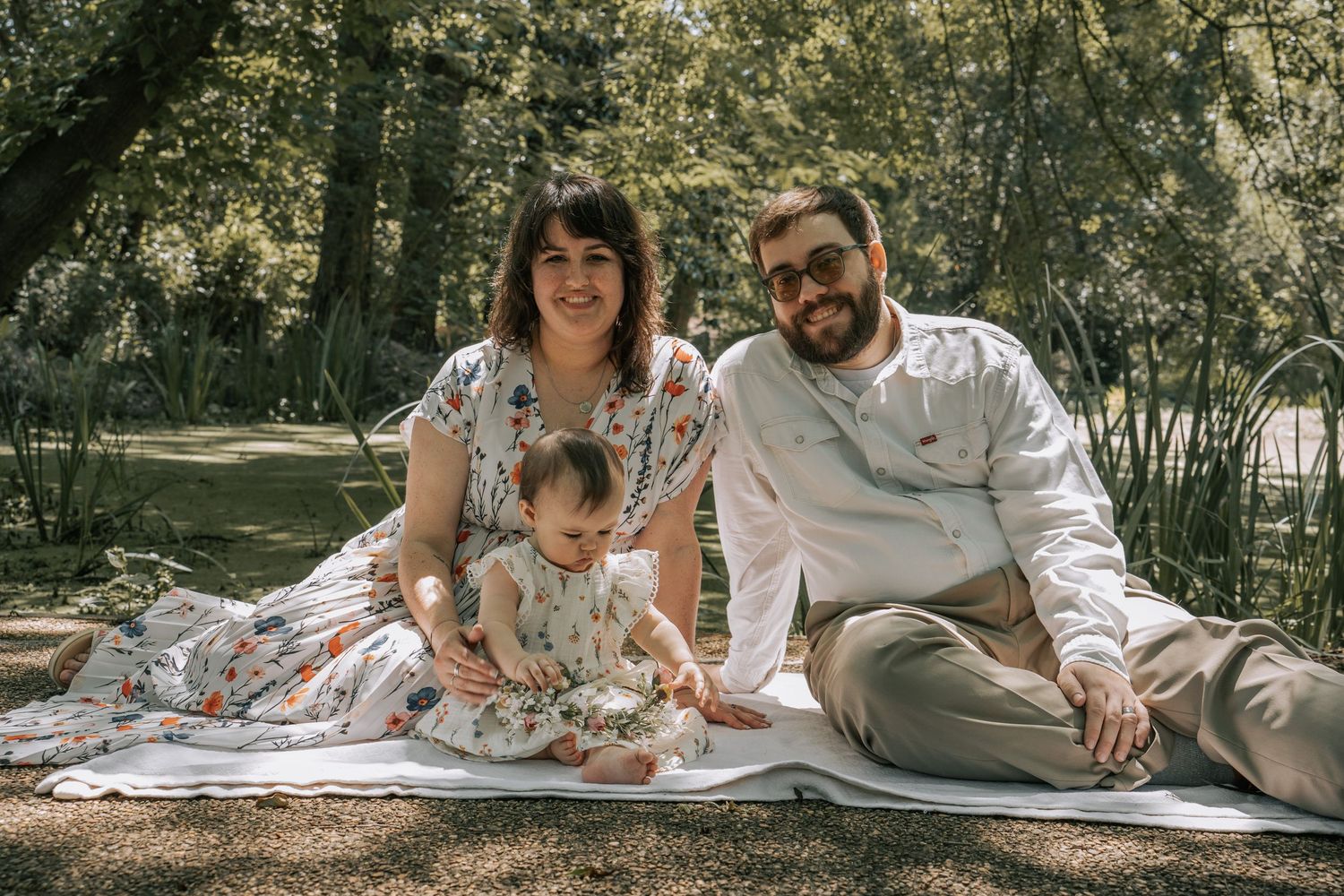 Happy family with baby enjoying a sunny outdoor picnic on a blanket surrounded by lush green trees.