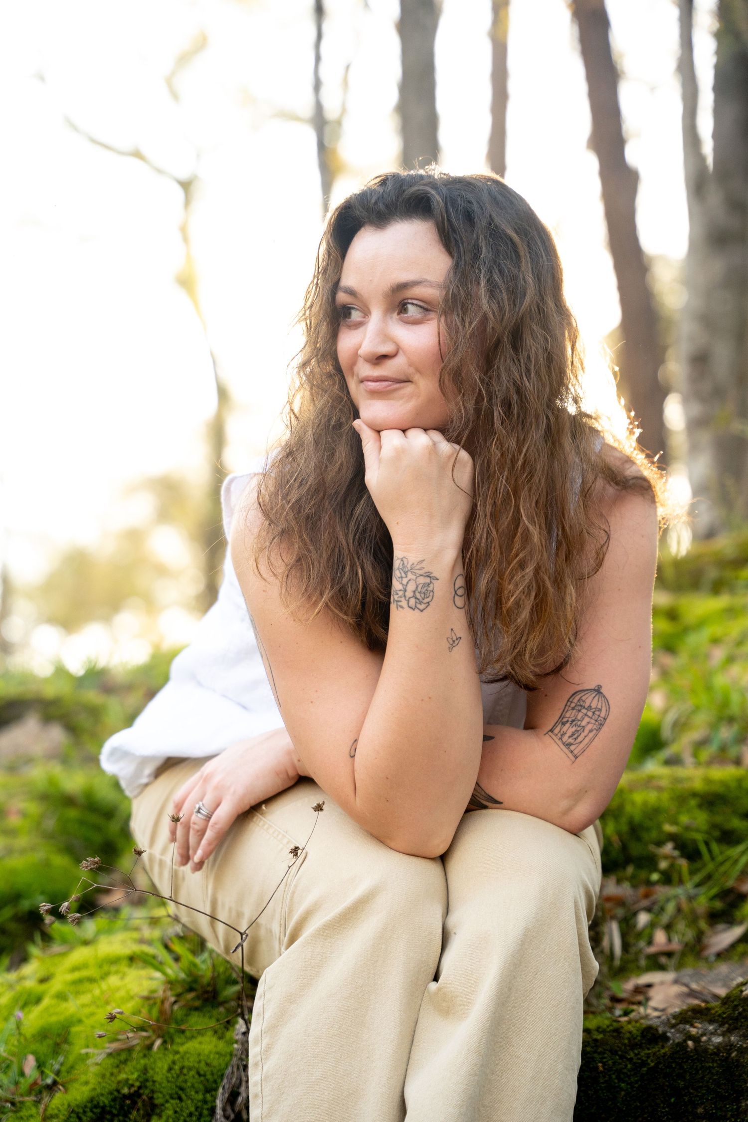 A woman with curly brown hair and tattoos sits thoughtfully in a sunlit forest, resting her chin on her hand.