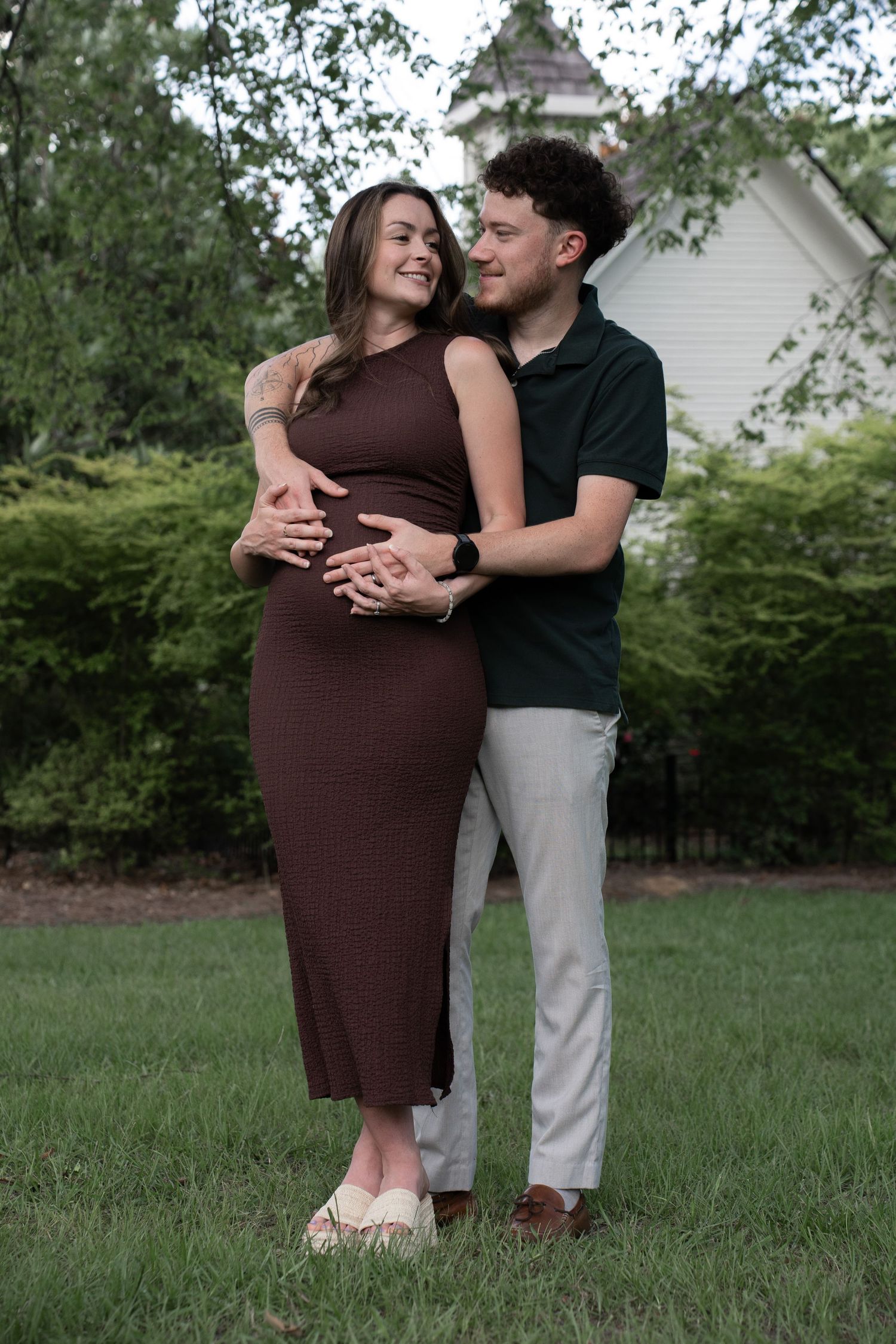 A couple shares a tender maternity moment outdoors, man embracing pregnant woman in brown dress amid lush greenery.