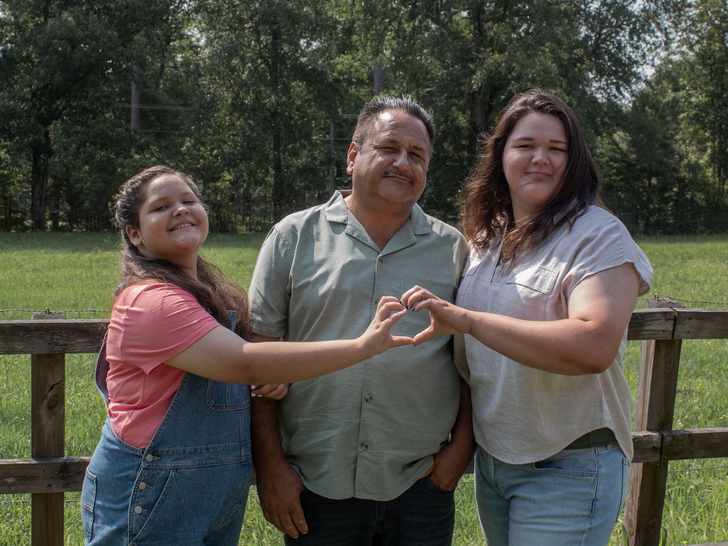 A man and two women pose outdoors near a wooden fence, forming a heart shape with their hands.