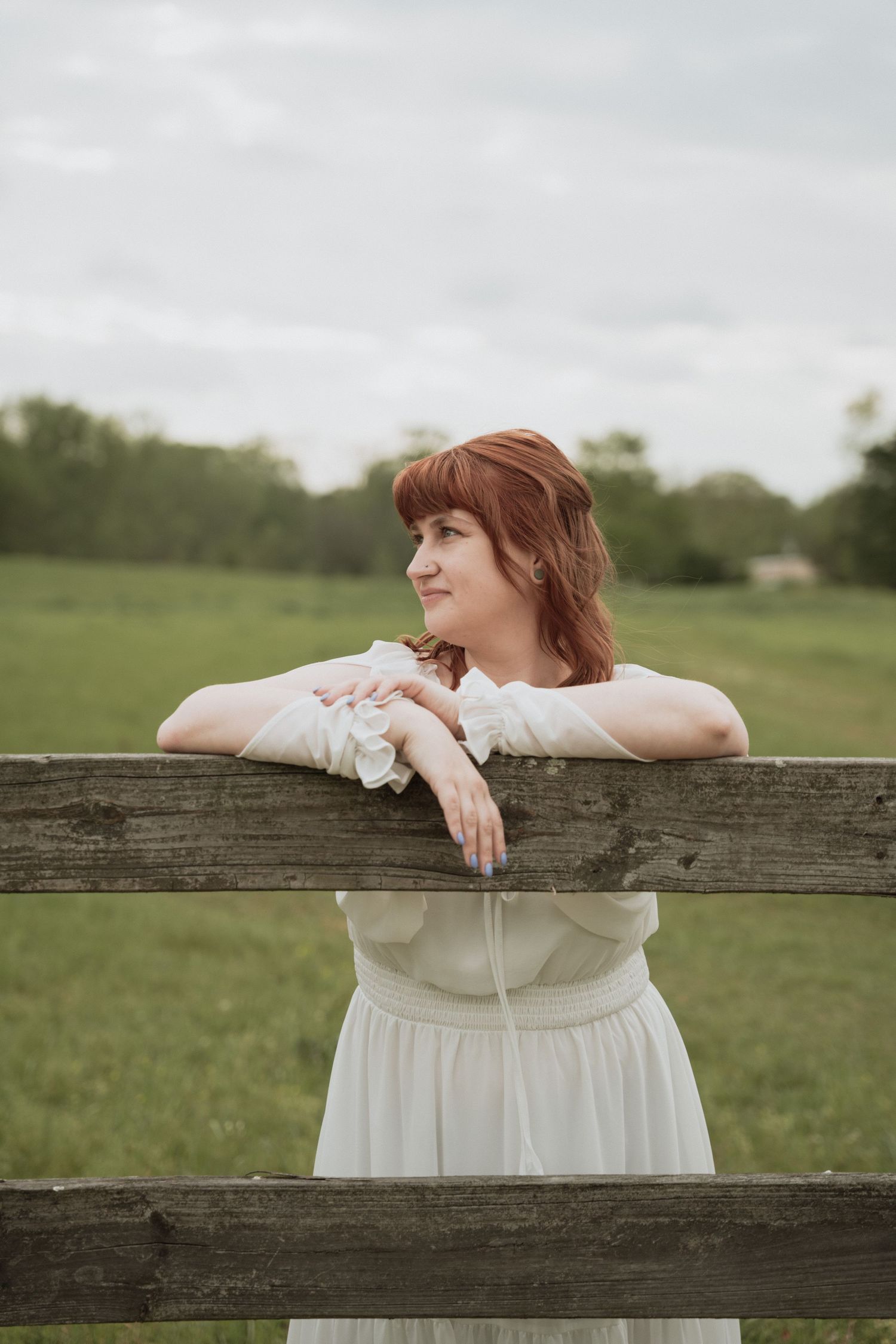 A young redhead woman in a white dress leans on a rustic wooden fence in a green countryside field.