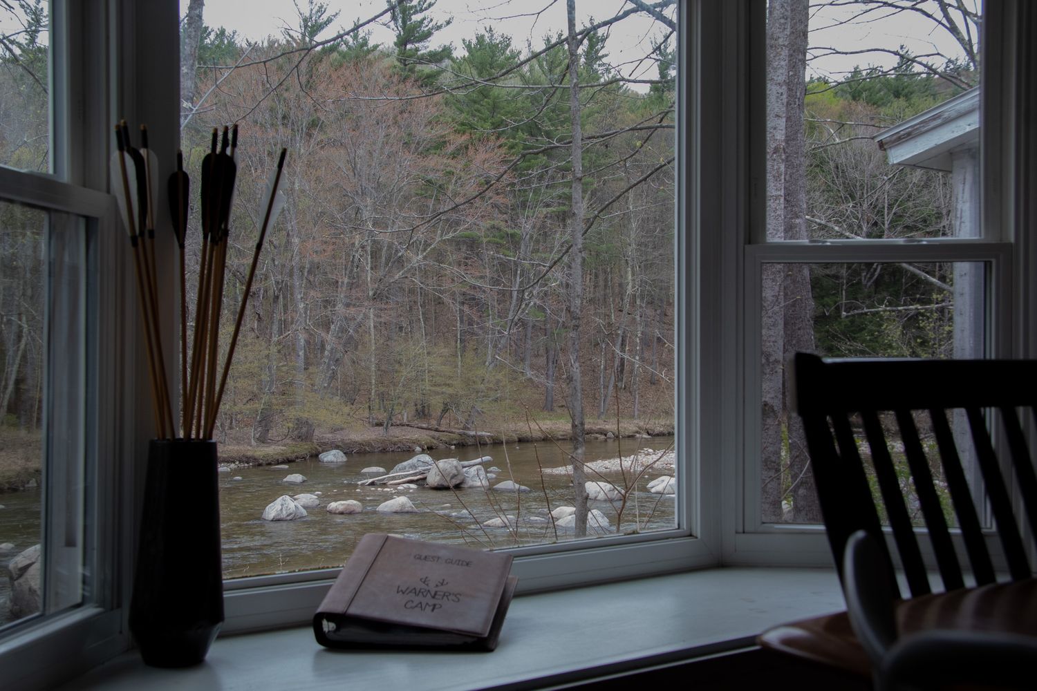 A cozy window sill with an open book and decorative sticks in a vase overlooking a serene forest stream.