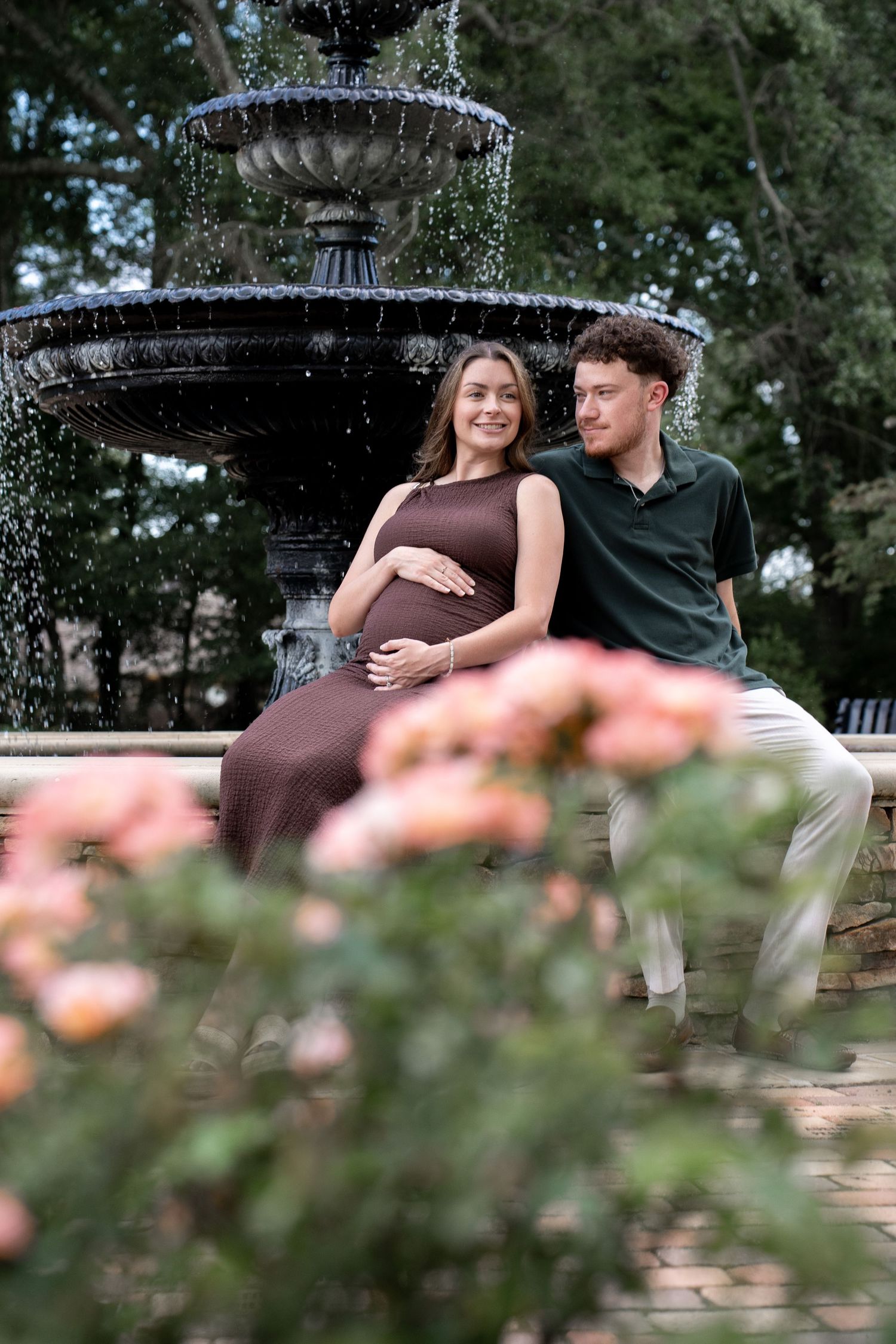 Pregnant couple sitting by ornate fountain with pink roses, woman in brown dress cradling baby bump, man in green polo.