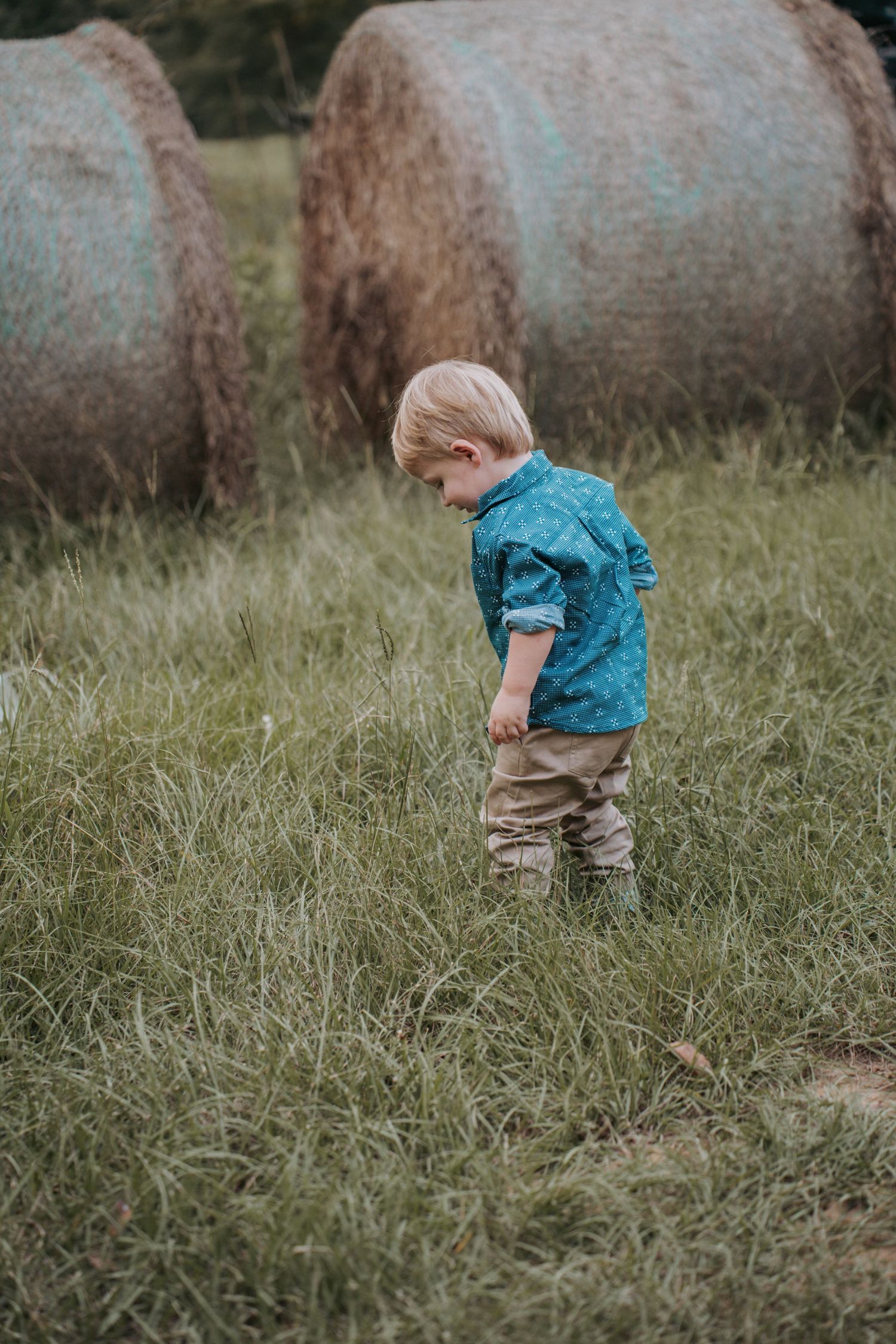A young blonde toddler boy in a teal shirt and tan pants walks through tall grass near large round hay bales.