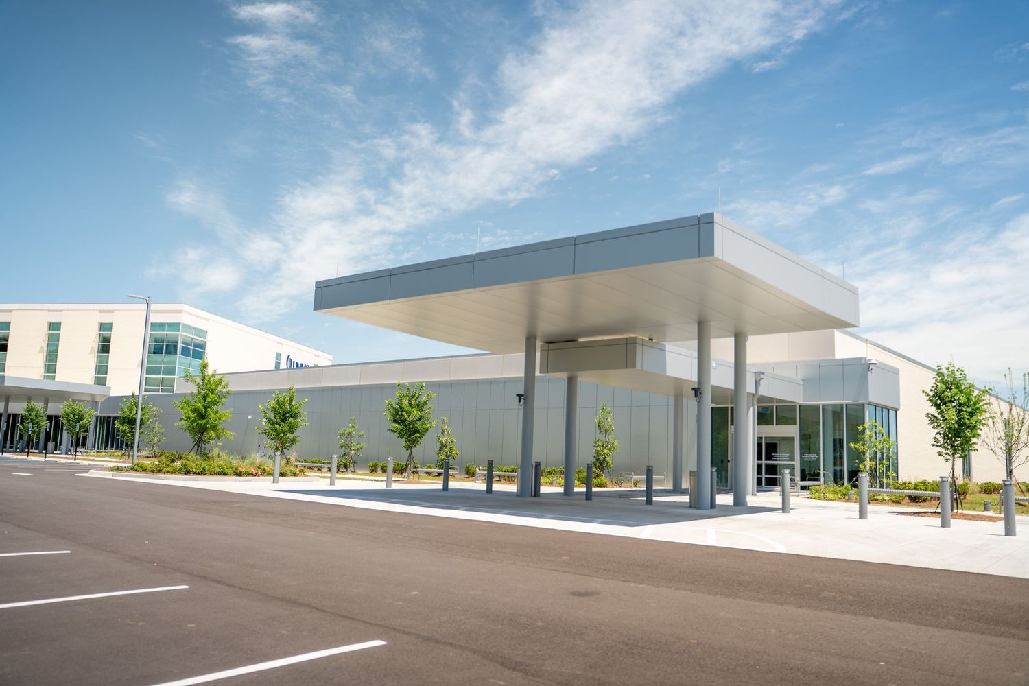 Modern commercial building with sleek canopy entrance, manicured trees, and spacious parking lot under a bright blue sky.