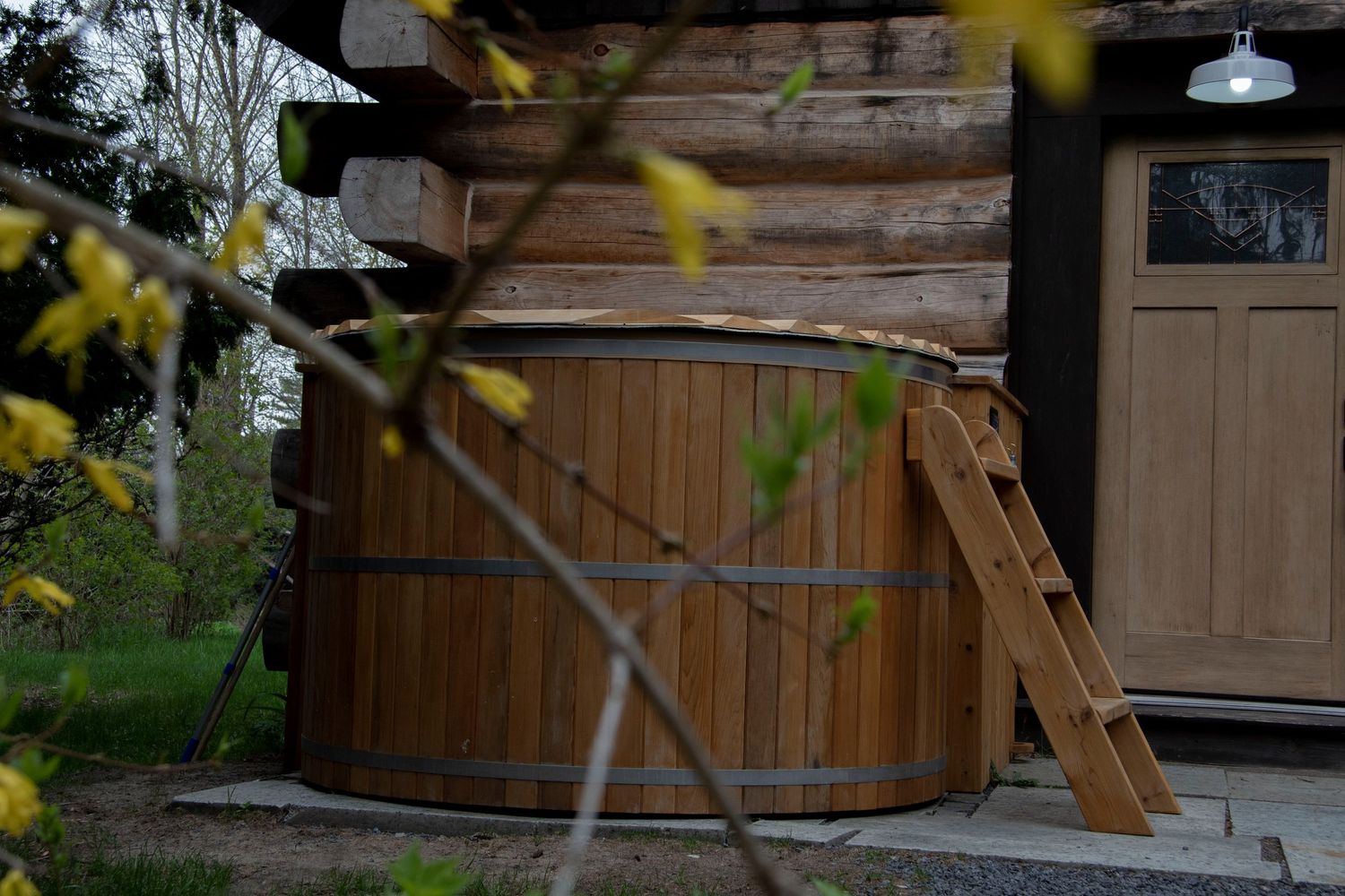 Rustic wooden hot tub with metal bands and ladder beside a log cabin, surrounded by spring foliage.