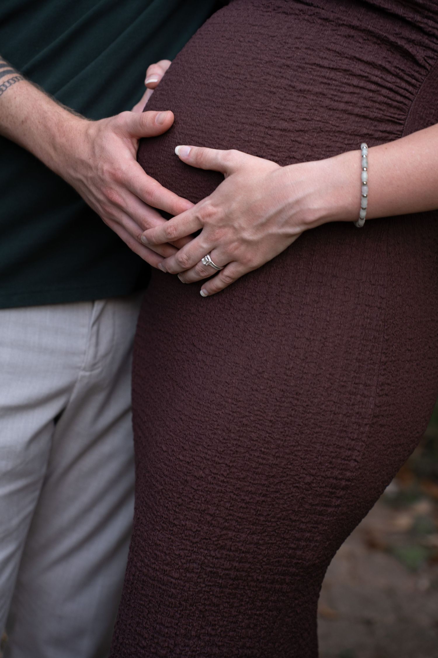 Couple tenderly cradling pregnant belly, woman wearing rings and bracelet, man in green shirt and gray pants.