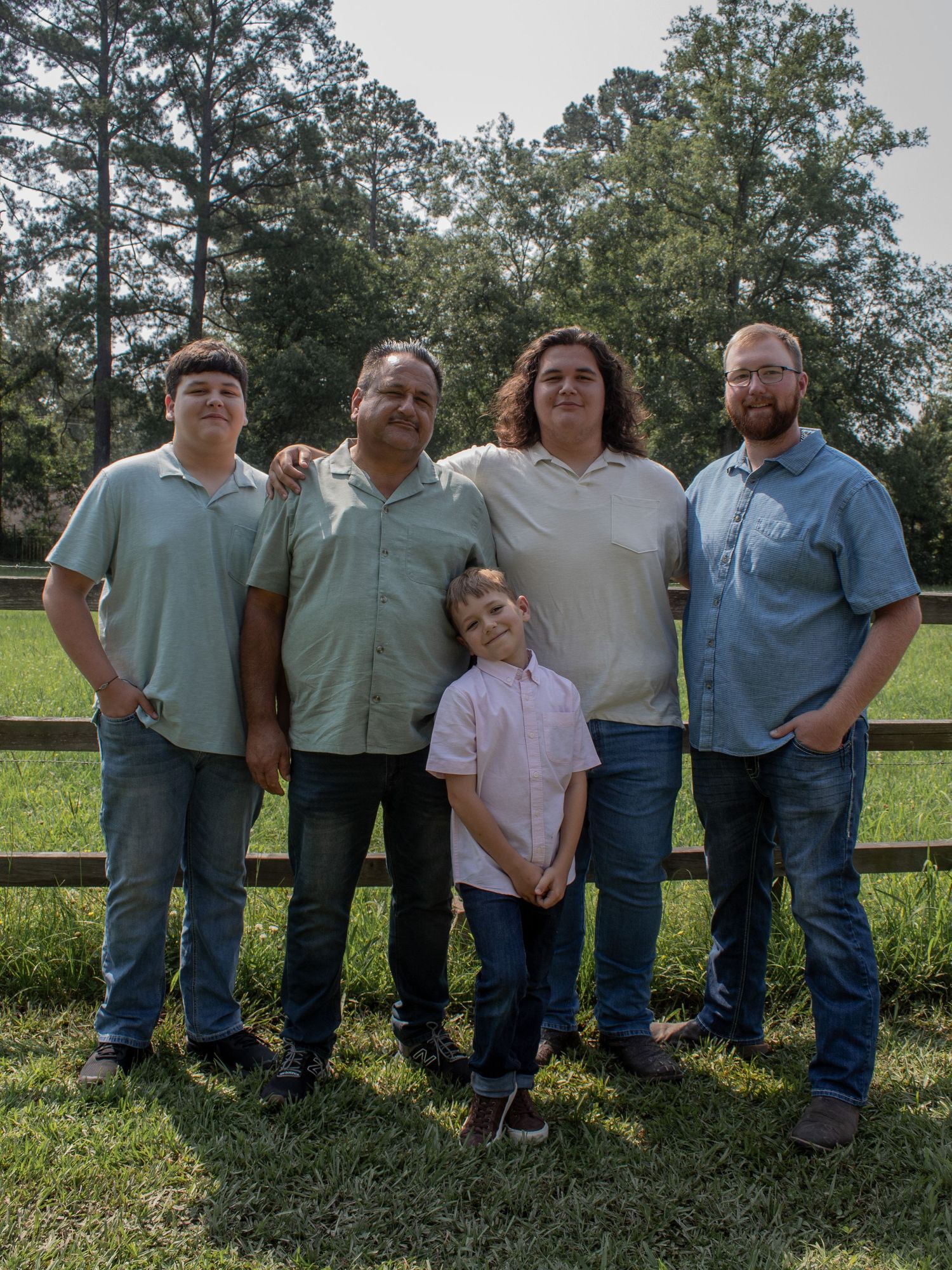 Family of five posing outdoors near a wooden fence on green grass, including four adults and one young child.