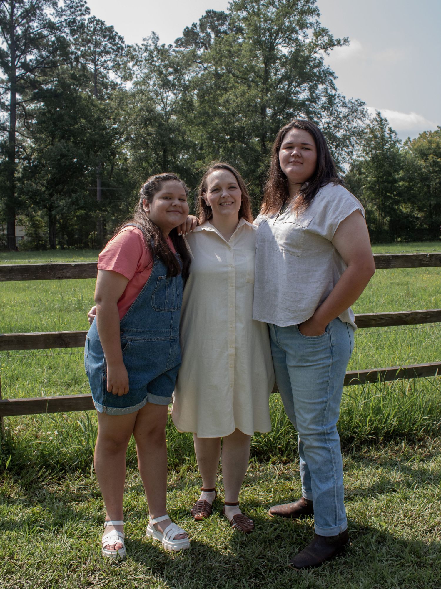 Three women smiling together outdoors near a wooden fence and green field on a sunny day.