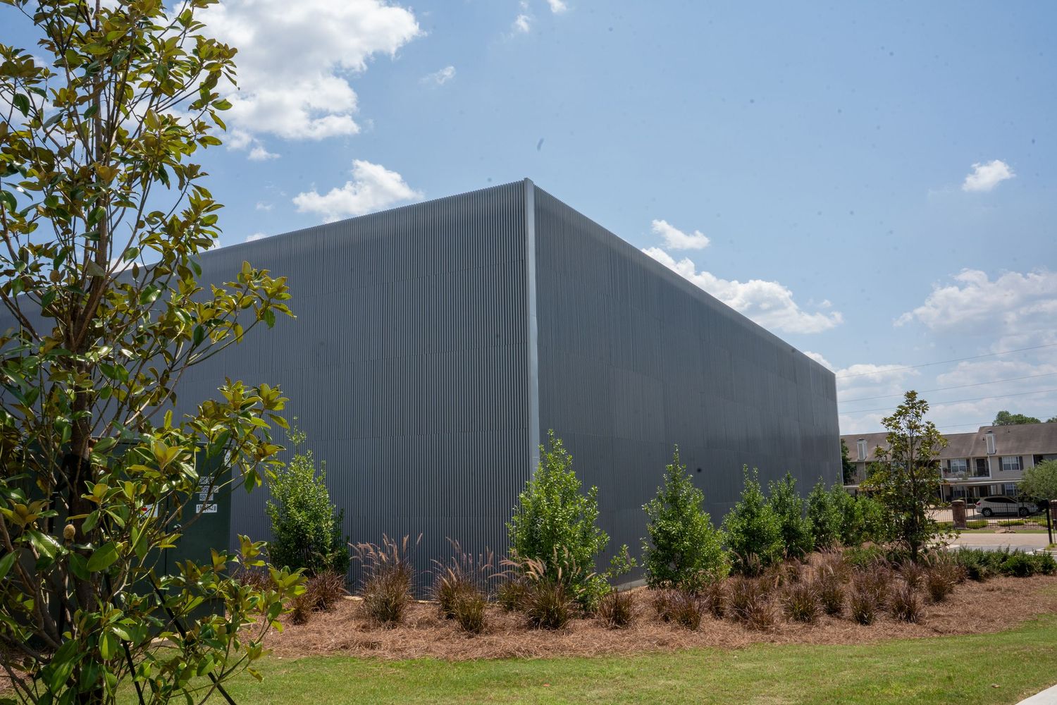 Large gray metal warehouse building surrounded by young evergreen trees and manicured landscaping under a partly cloudy sky.