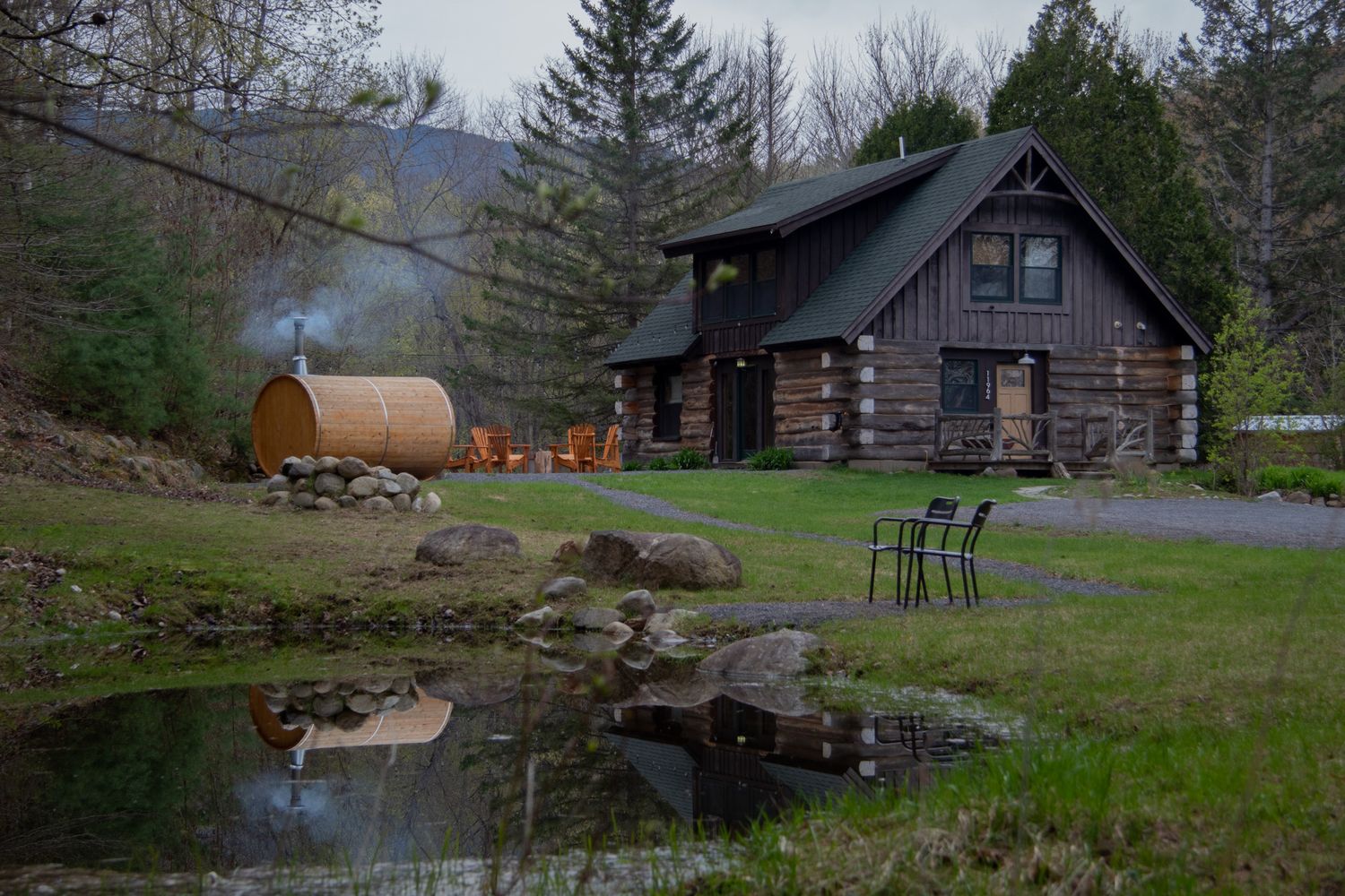 Rustic log cabin with barrel sauna, pond, and green chairs nestled in a lush mountain forest setting.