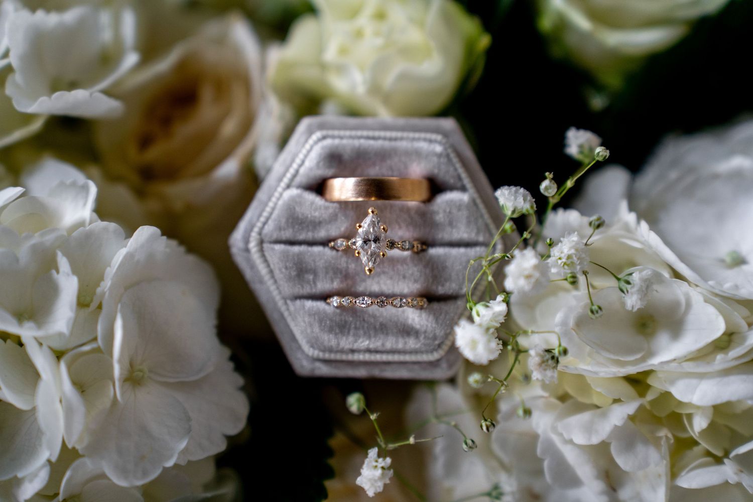 Gold wedding rings displayed in a velvet hexagon box surrounded by white hydrangeas and roses.