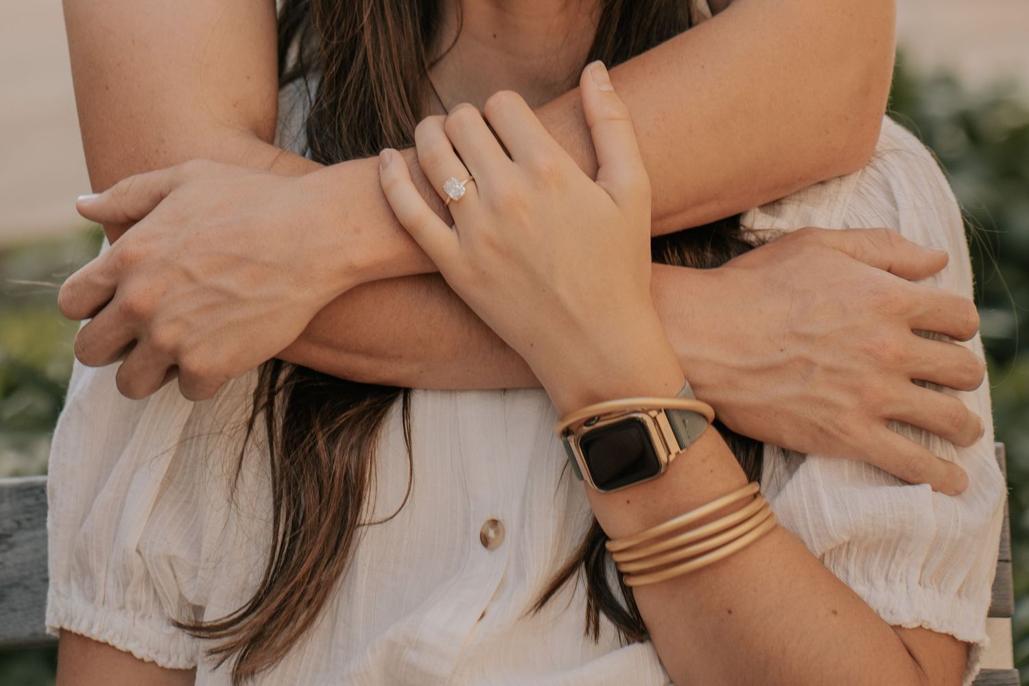 Two women embracing, wearing gold jewelry including a smartwatch band, bracelet, and ring, dressed in white.