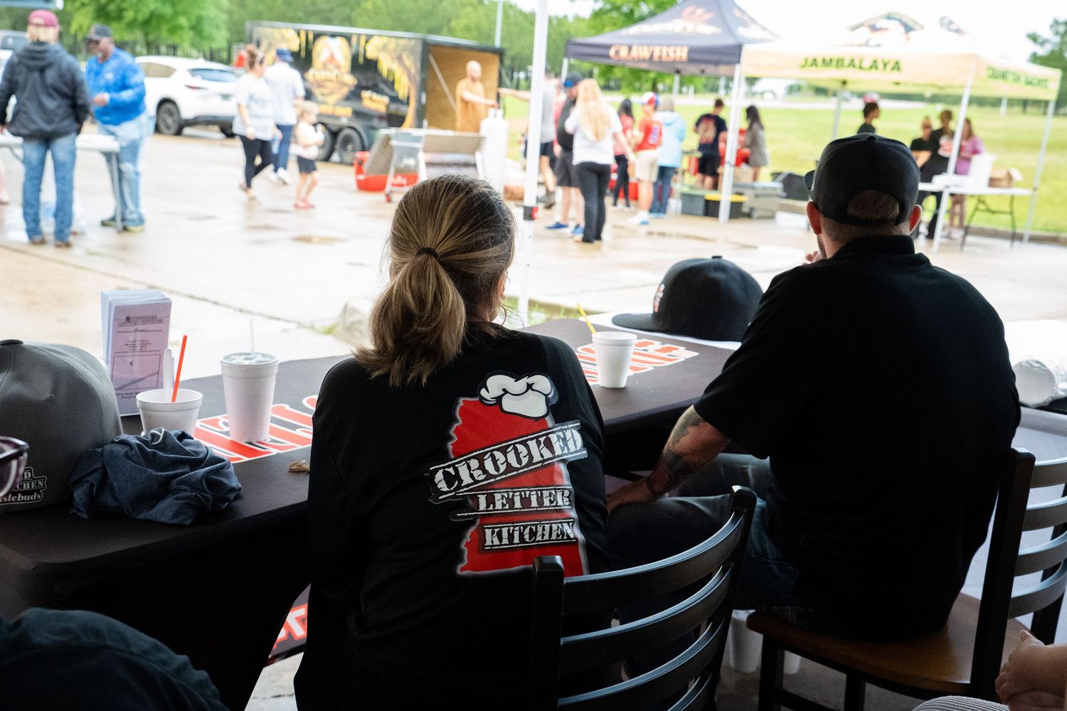 Outdoor festival crowd with vendors, featuring a woman in a Crooked Letter Kitchen shirt seated at a table.