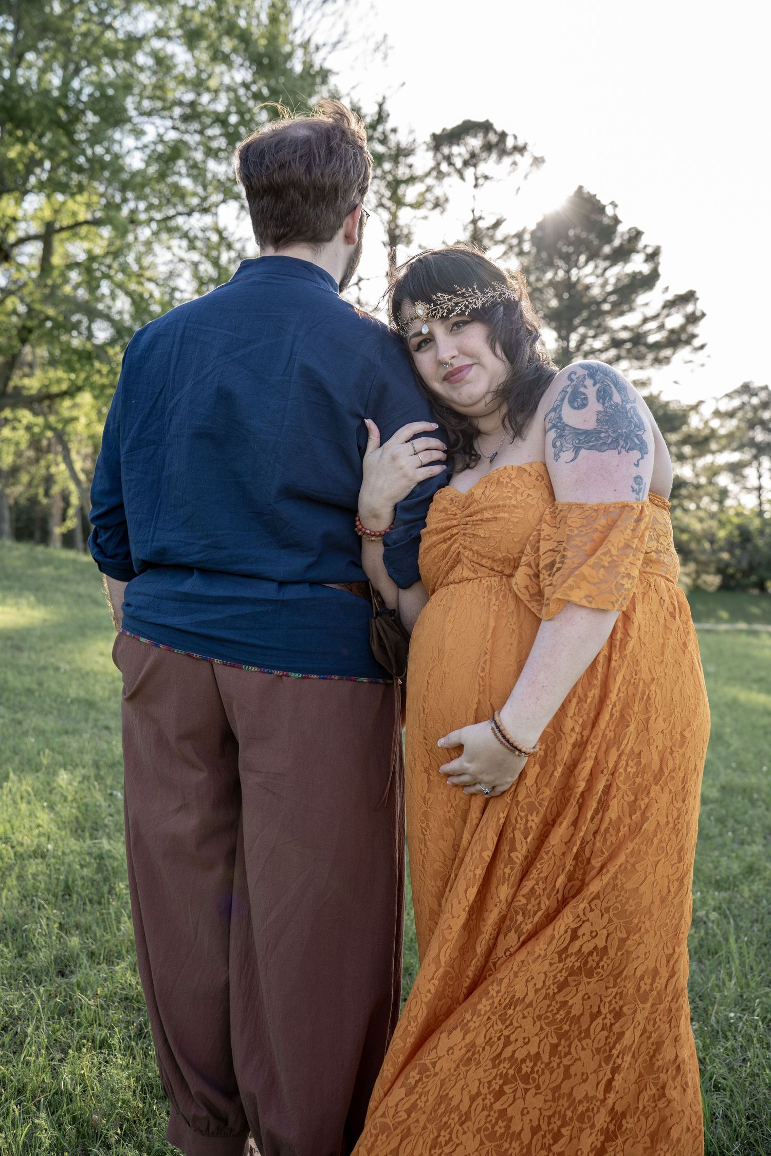 Pregnant woman in orange dress with tattoos embraces partner outdoors in green park during golden hour maternity photoshoot.