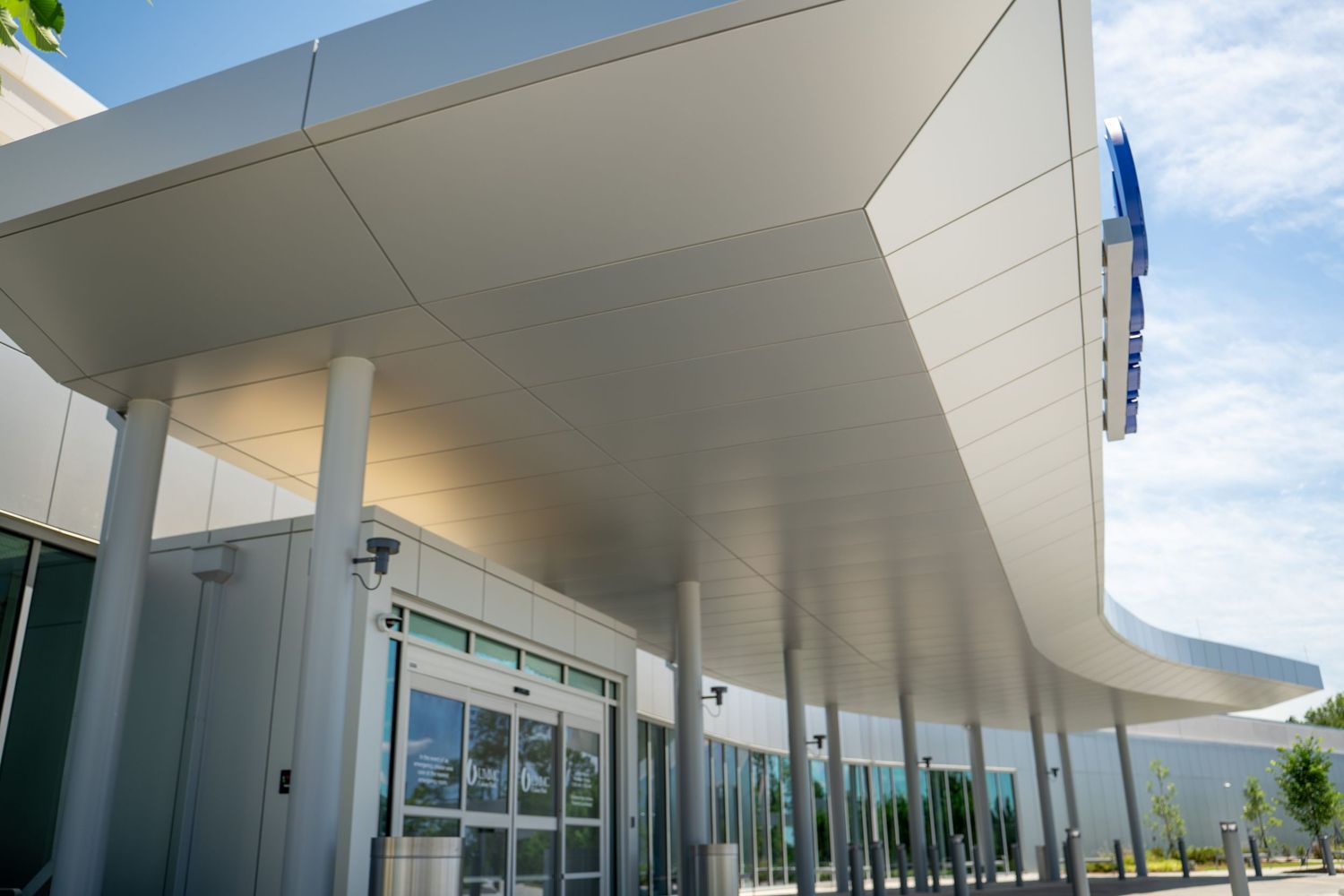 Modern building entrance with sleek white canopy, glass automatic doors, and blue signage under a clear sky.