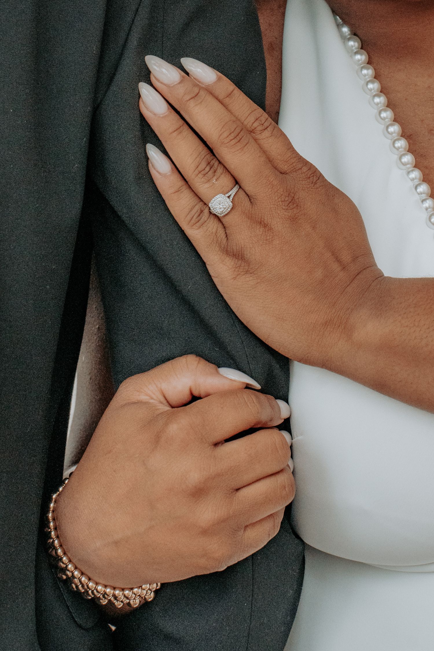 Close-up of bride's hand with diamond halo engagement ring and pearl bracelet resting on groom's dark suit jacket.