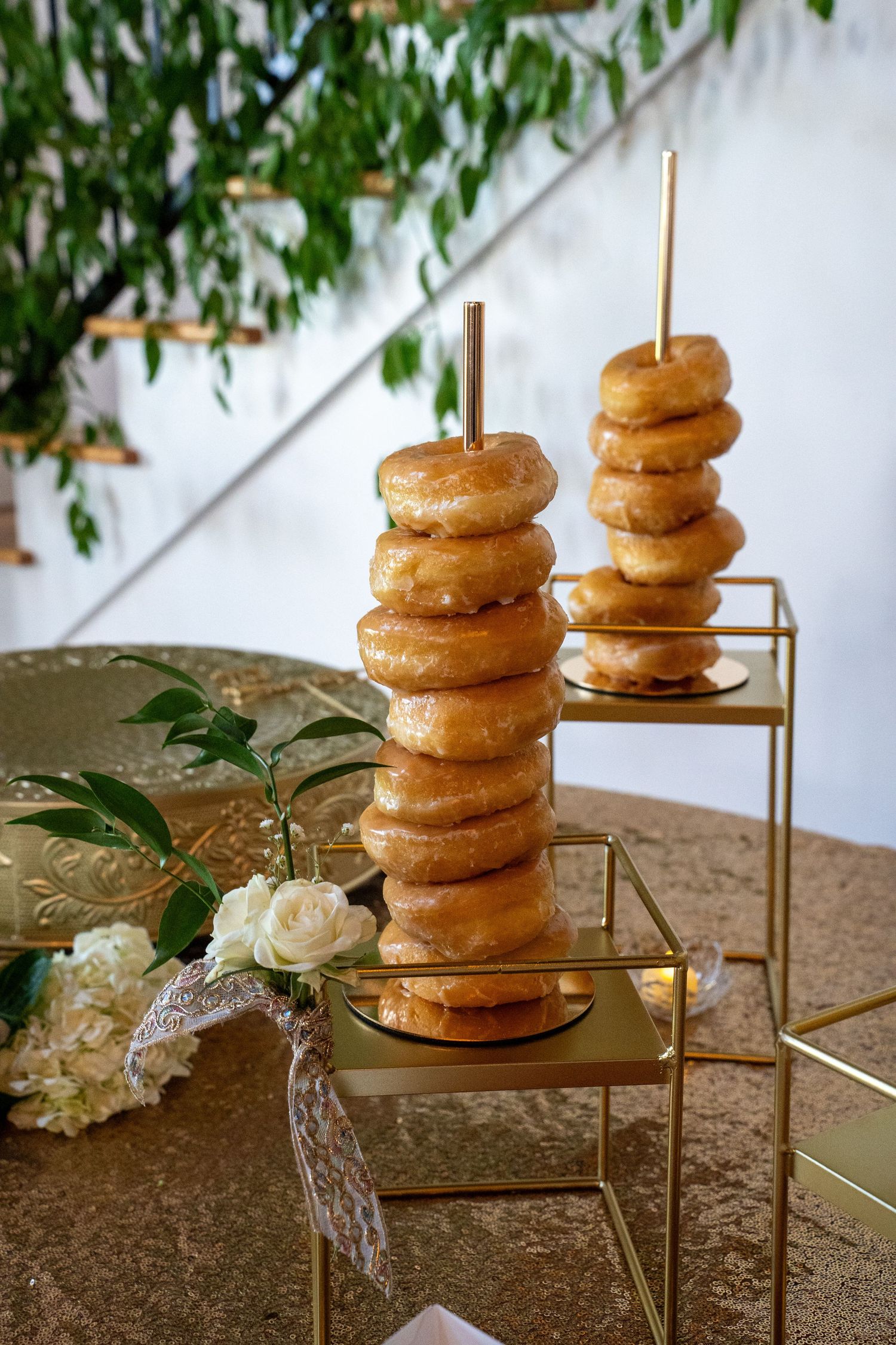 Glazed donuts stacked on gold rod stands displayed on a gold glitter tablecloth with white floral arrangements.
