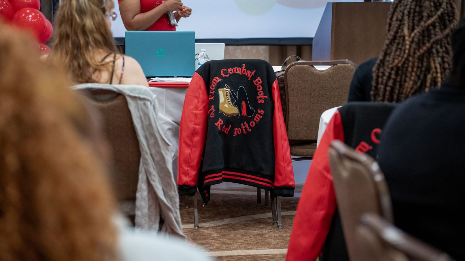 Red and black varsity jacket on chair at indoor event with attendees in background.